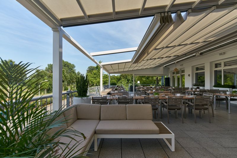 Reference picture of a pergola stretch with cream fabric cover and white frame over the outdoor area of the "bootshaus" in Mannheim. On the picture the awning is about to retract